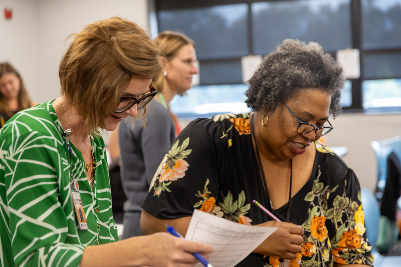 Two educators stand and talk while holding papers and pens.
