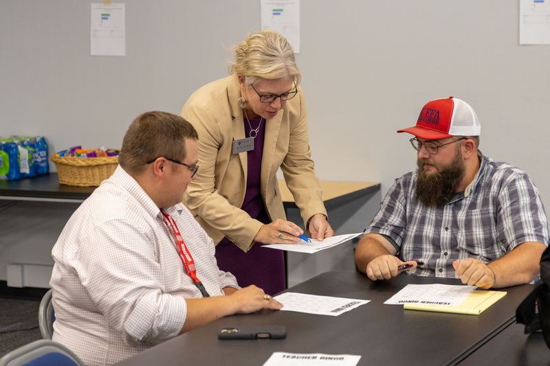 An educator stands between two other educators who are seated. She is leaning down pointing to her paper with a pen.