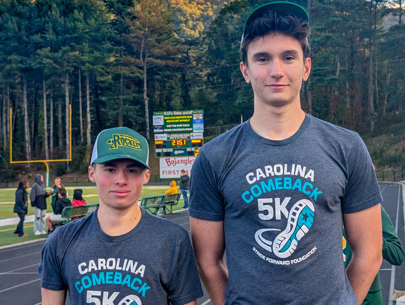Pablo and Matthew stand in front of the Reynolds scoreboard wearing A.C. Reynolds hats and Carolina Comeback t-shirts