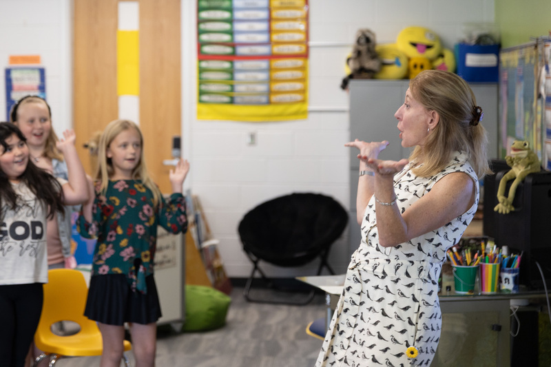 A close-up shot of Ms. Melody standing in front of the room, facing her students who are holding up their hands as they sing a choreographed song.