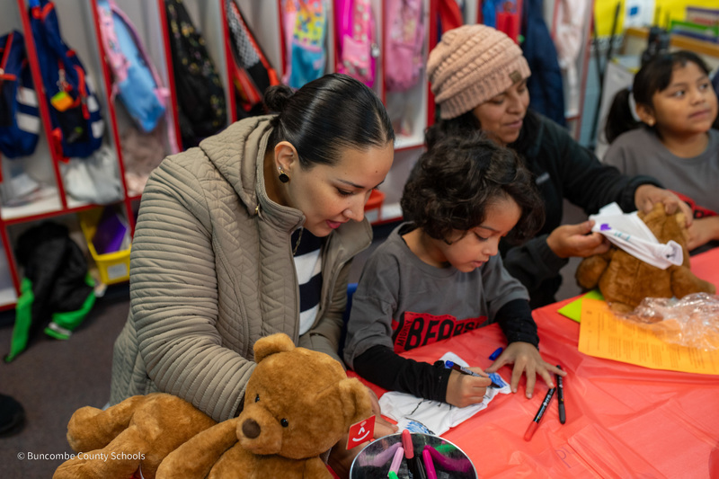 Parents sitting with students and their teddy bears help students color a headband with a bear design.
