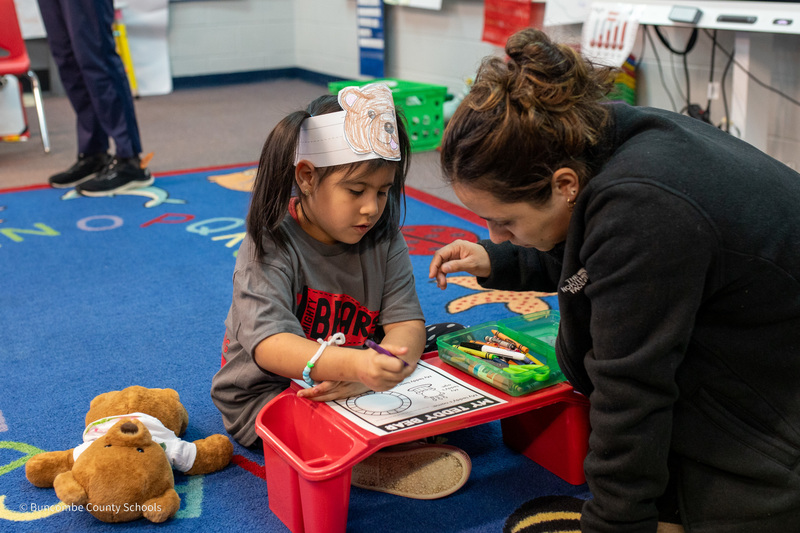 Kendra and her mom sit on the floor with her teddy bear. Kendra is coloring on a worksheet.