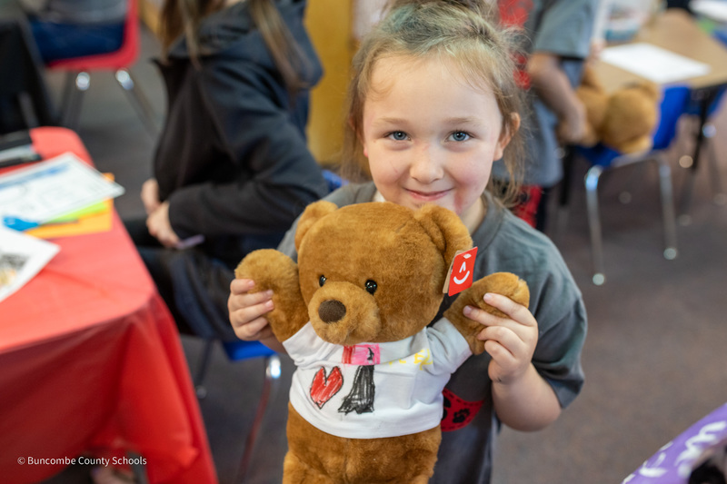 A student smiles as she stands and holds her teddy bear.