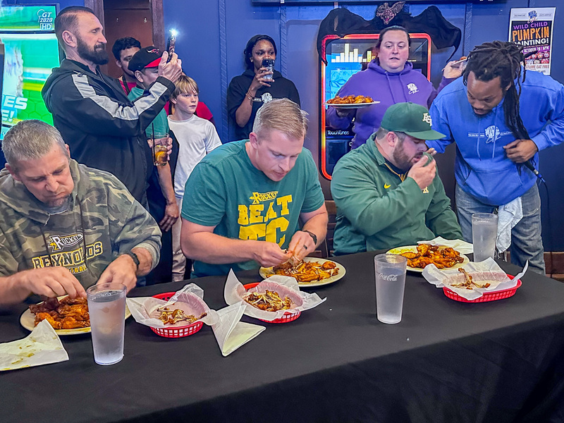 Mr. Bowling, Coach Greer, and Coach Staicar at the Reynolds team table eating from plates of wings. They are baskets for bones in front of them.