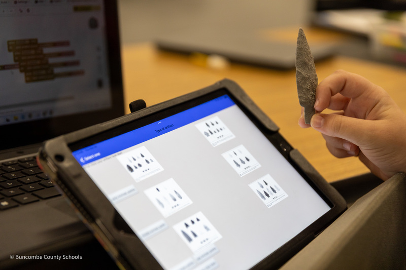 A closeup of a table displaying the archaeological app, and a hand holding an arrowhead next to the tablet.