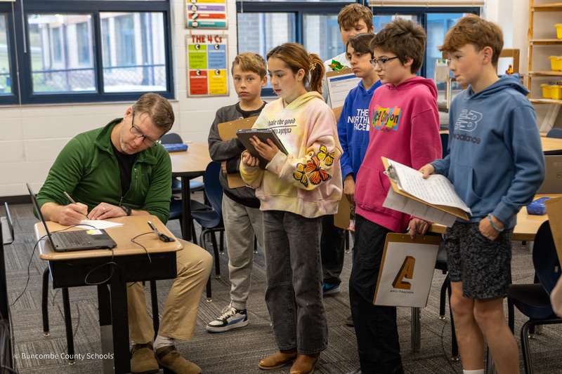 Mr. Thompson sits at a desk as students present their projects.