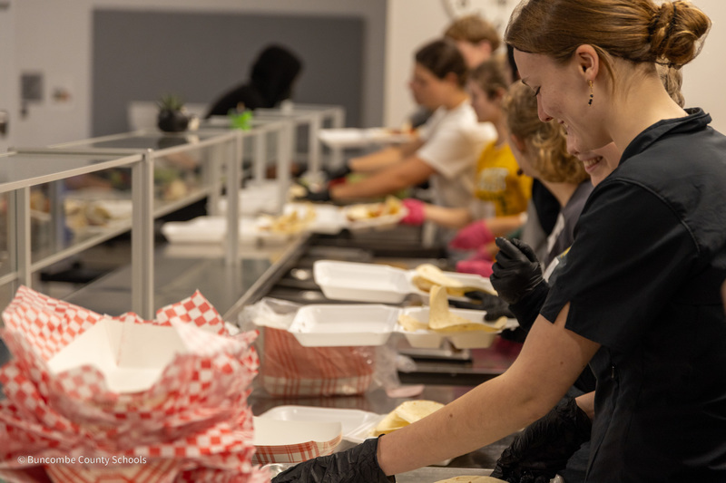 Students behind the serving line at Transformation Village dish food into containers