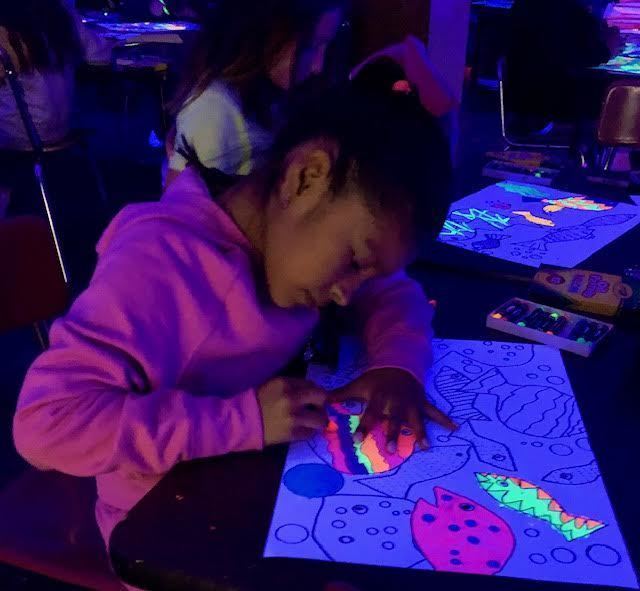 Dark image of a student using florescent paint to make an ocean scene under a black light.