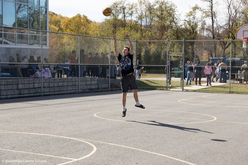 Male student jumping in the air on the basketball court as he is shooting the basketball toward the goal.