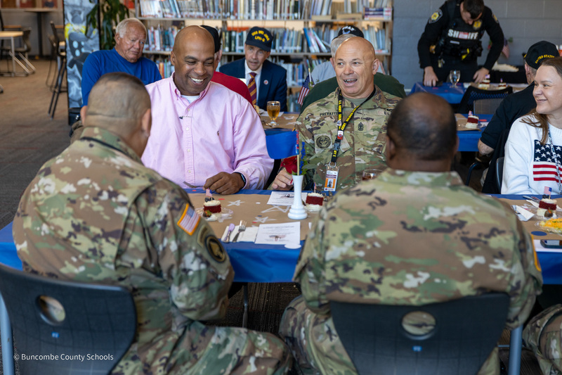 Principal Stanley Wheeless sits at a table with Army personnel in camo fatigues
