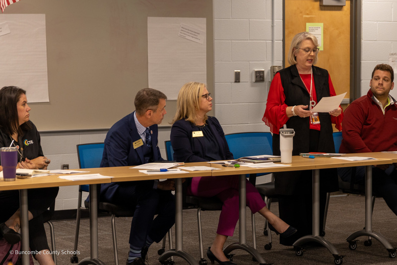 A woman stands next to people seated on a panel. She is reading from a sheet of paper.