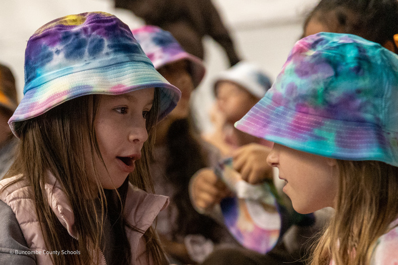 Two students wearing tie-dyed thinking caps turn and talk to each other.