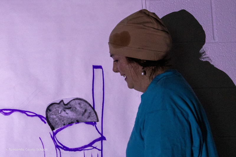 A teacher dressed as Harold looks at her drawing of a bed on the wall.
