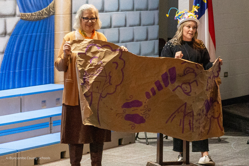 Two teachers stand in the front of the theater holding a giant map