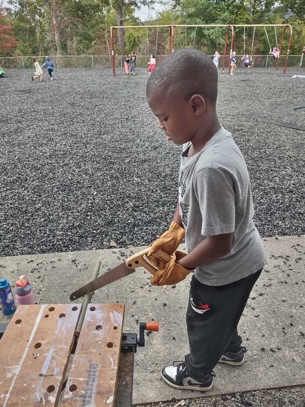 Little boy in gray shirt and black pants, wearing safety gloves, using a hand saw to cut a small piece of bamboo.