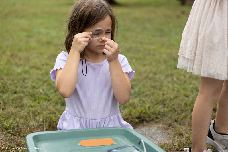 Little girl in purple dress using a small magnifying glass to look at something in her hand.