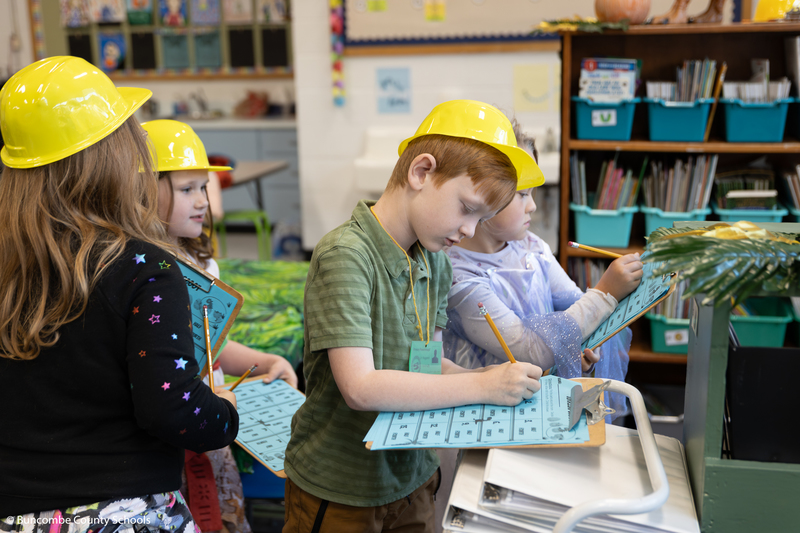 Group of students standing with a clipboard, and sheet of paper. One boy is writing on his piece of paper.