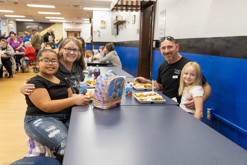 Two adults eat with students at cafeteria tables at Leicester Elementary