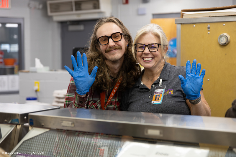 A nutrition volunteer stands next to BCS Associate Superintendent Dr. Jennifer Reed in the serving line. They are wearing safety gloves and waving.
