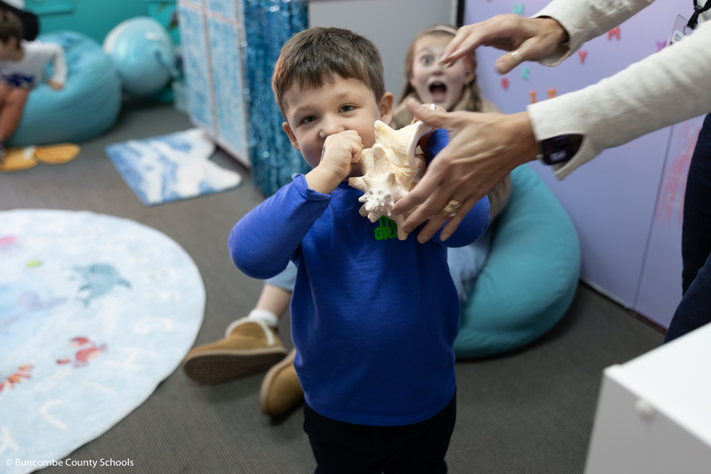 Little boy holding up a sea shell to his ear, listening to see if he hears the ocean in it.