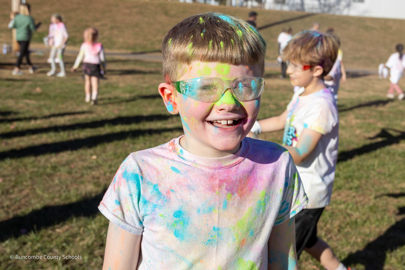 A student poses for a photo. His hair, safety glasses, and shirt are covered in colorful powder