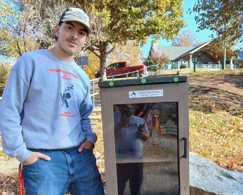 A student stands next to a "little free library" box he has built at the end of a driveway.
