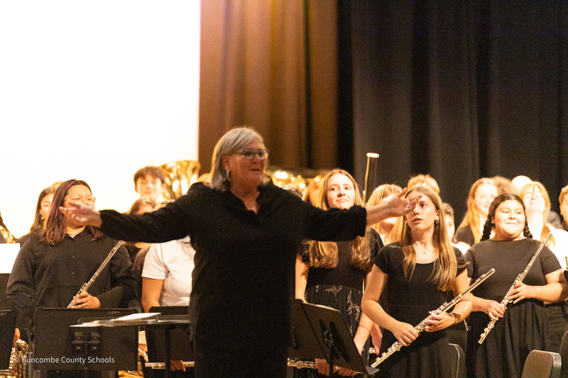 A clinician smiles and holds her arms out in front of student musicians. They are standing with their instruments for an ovation.