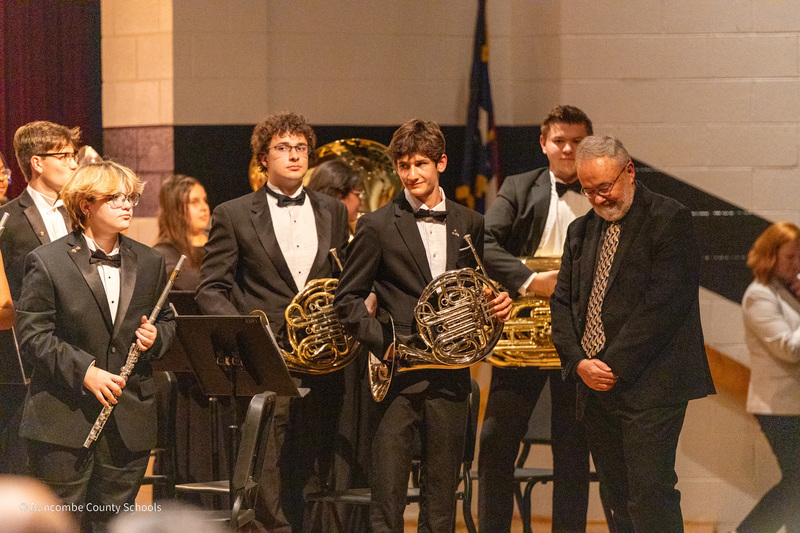A clinician stands next to student musicians on stage. They are standing, holding their instruments, for an ovation.