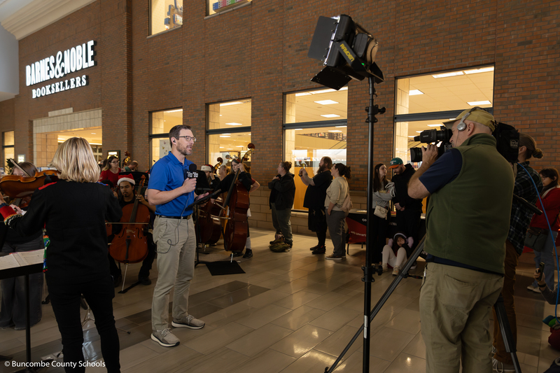 WLOS meteorologist Jason Boyer speaking on camera in front of the orchestra as they perform.