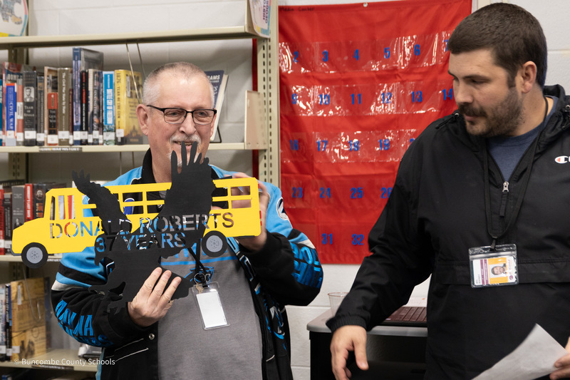 Mr. Roberts stands next to Mr. Maney and holds a metal Blackhawk school bus wall hanging that has his name on it.