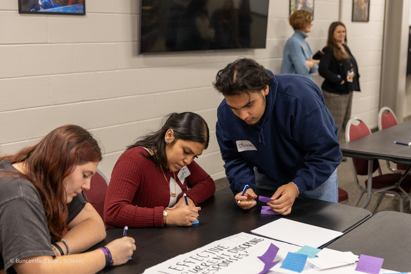 Three students - two seated and one standing - write on post-it notes at a table.