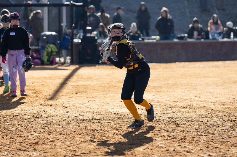 Girl wearing black and yellow uniform running and getting ready to throw a softball.