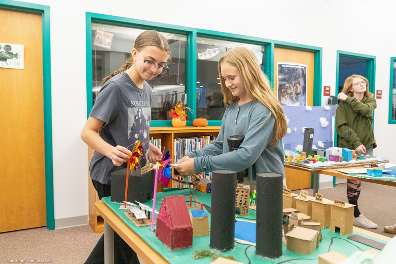 Two students adjust their diorama while smiling.