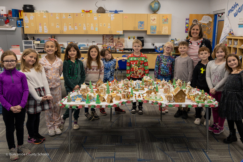 Mrs. Gortney and her class stand behind a table filled with their gingerbread creations