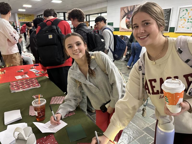 Two girls smiling at the camera and writing on their holiday card. 