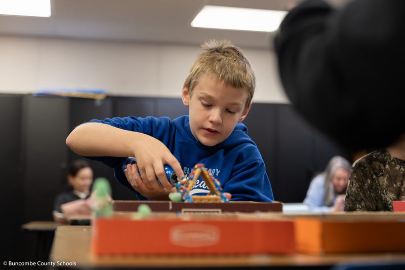 Little boy in blue hoodie applying icing to his gingerbread house.