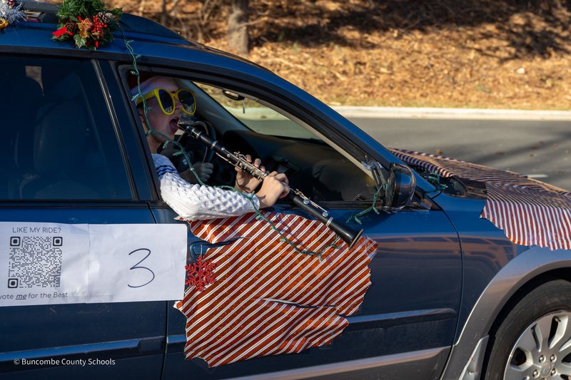 A band student wearing sunglasses and riding in the passenger seat of a car plays his clarinet out the window