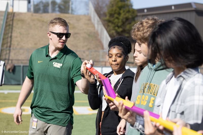 Reynolds teacher Chandler Greer stands outside with his class. Students are holding colorful plastic tubes to form a chute. He is dropping a golf ball at the beginning.