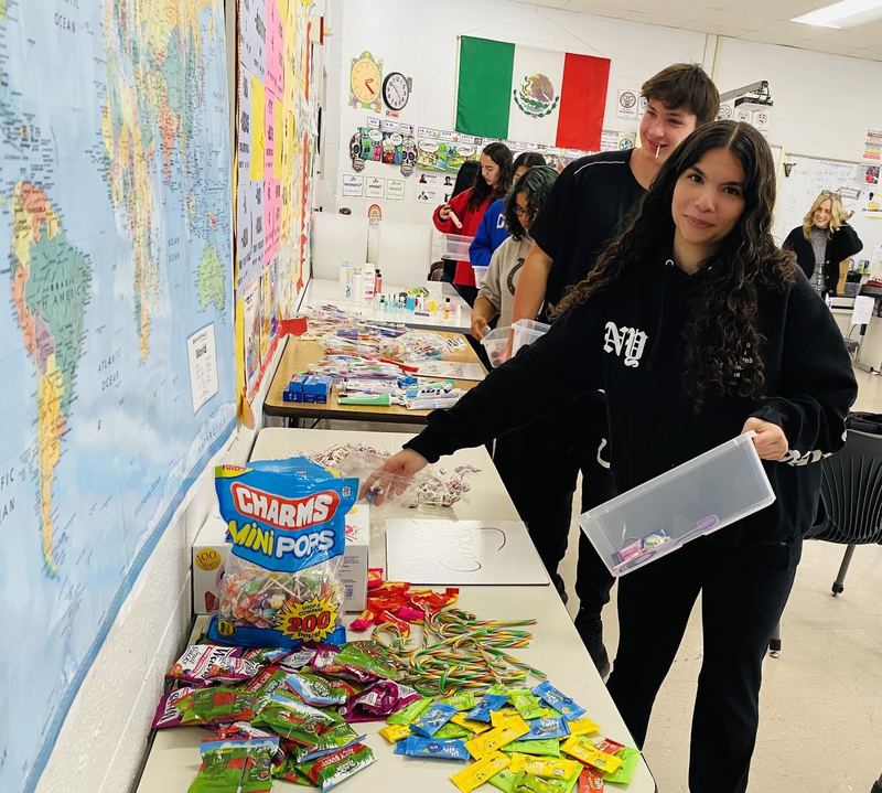 Photo of students walking beside tables lined with goodies while carrying a box and filling it with items. 