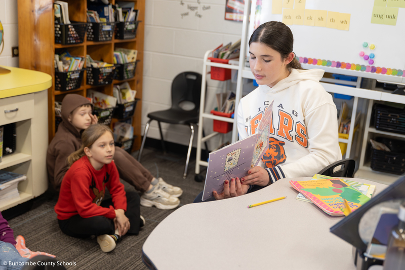 Female student reading a book to a small group of third graders. 