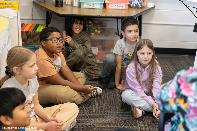 Group of third grade students sitting in the floor listening to a middle school student read a book. 