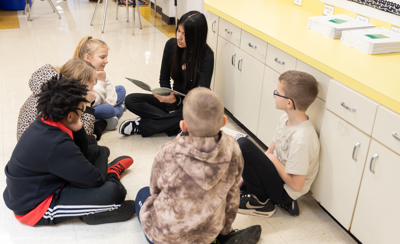 Group of students sitting on the floor listening intently to a middle school girl reading a book. 