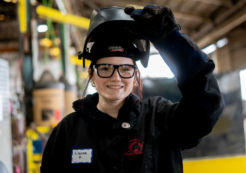 A student raises her welding mask and smiles