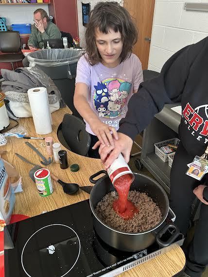 Female student purring an open can of tomatos into a pot with browned ground beef as they make chili.