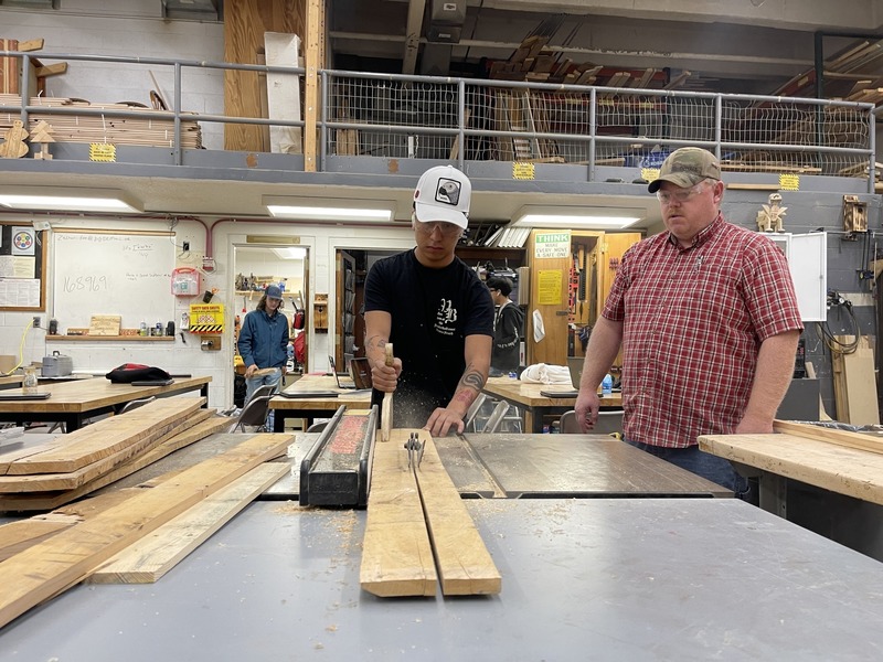 A teacher watches a sudent using a circular saw in the school wood shop.