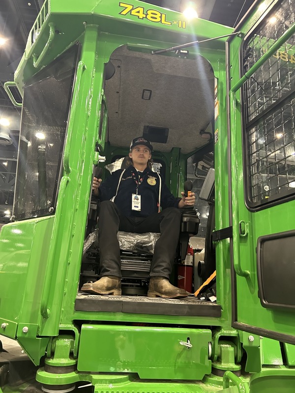 A student dressed in an FFA jacket operators a tractor.