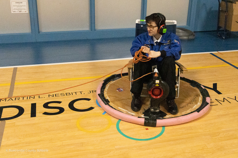 A student is seated on a student-built hovercraft on the Nesbitt gym floor.