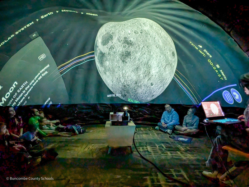 People sit inside a geodome with a moon projected on its wall.