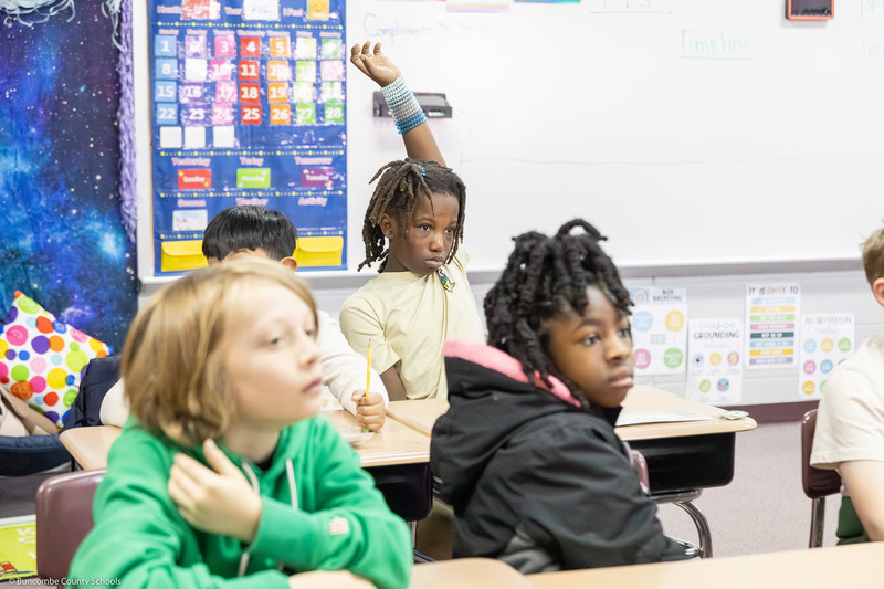 A student raises his hand among a row of desks.