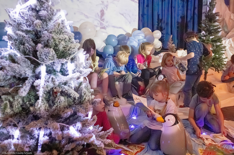 A group of students sit and read in the middle of a winter wonderland scene featuring Christmas trees, penguins, and snow props.